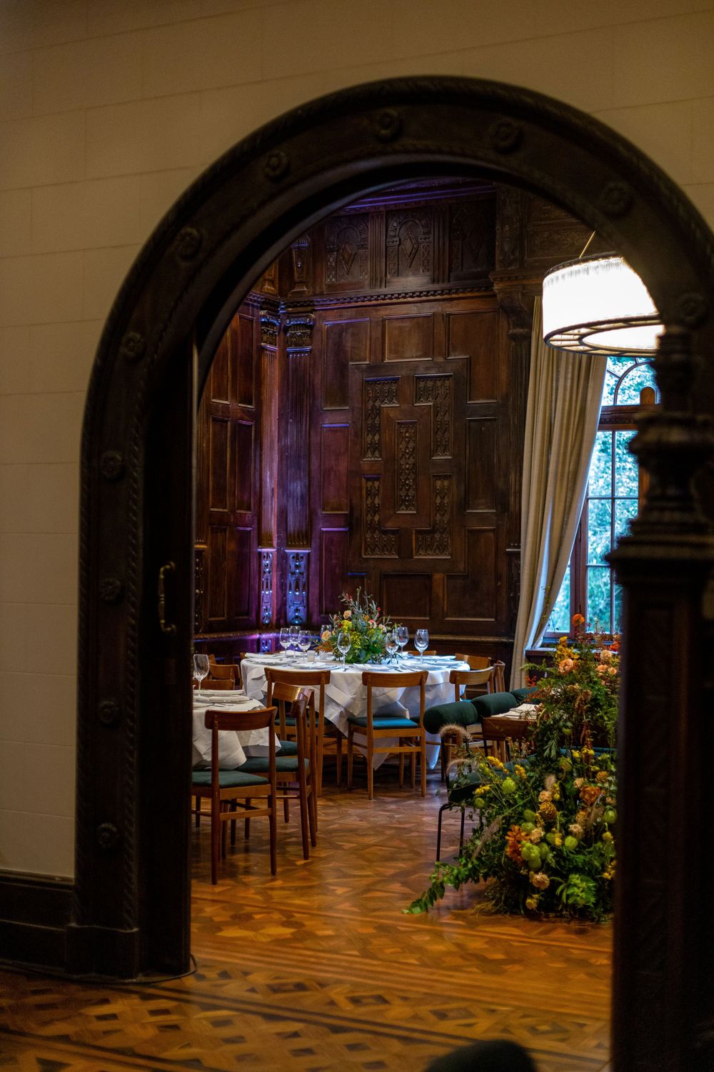 Elegant dining room with dark wood paneling viewed through an arched doorway with floral arrangements and table settings.