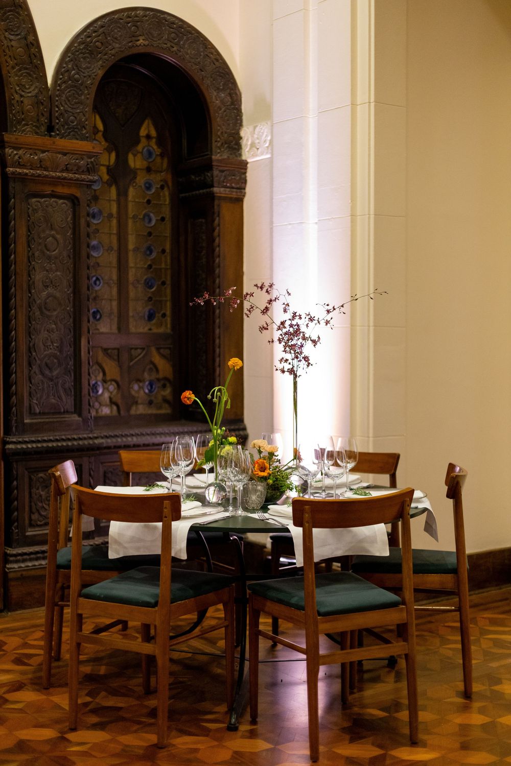 Elegant dining room interior with wooden tables, green chairs, and ornate carved wood panels in warm lighting.