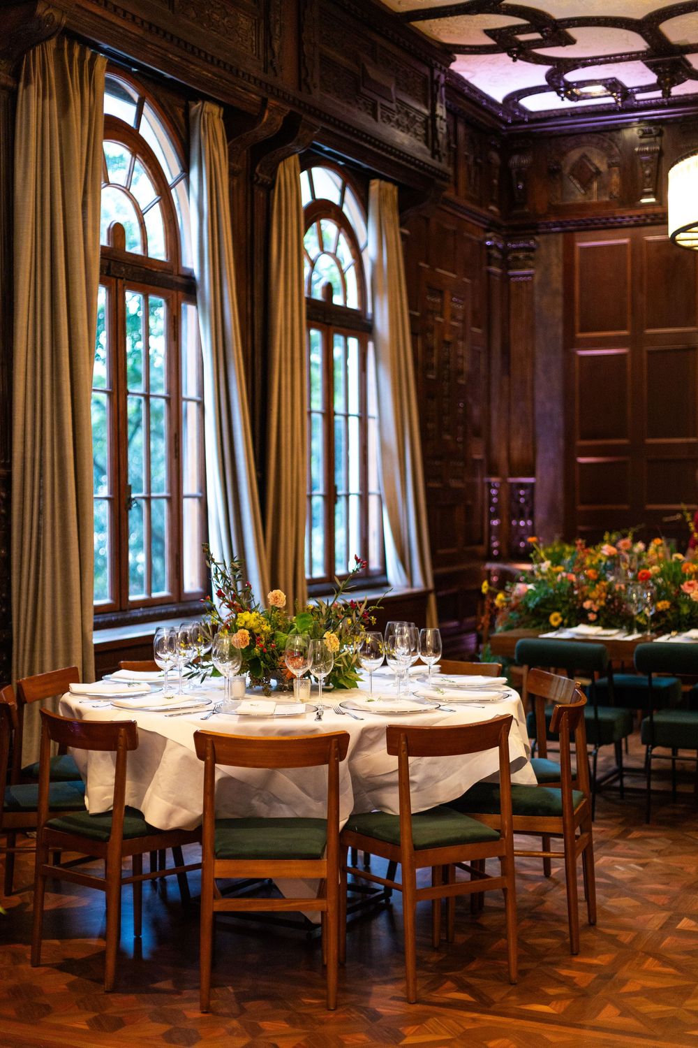 Elegant dining room with wood-paneled walls, arched windows, and tables set with white linens and floral centerpieces.