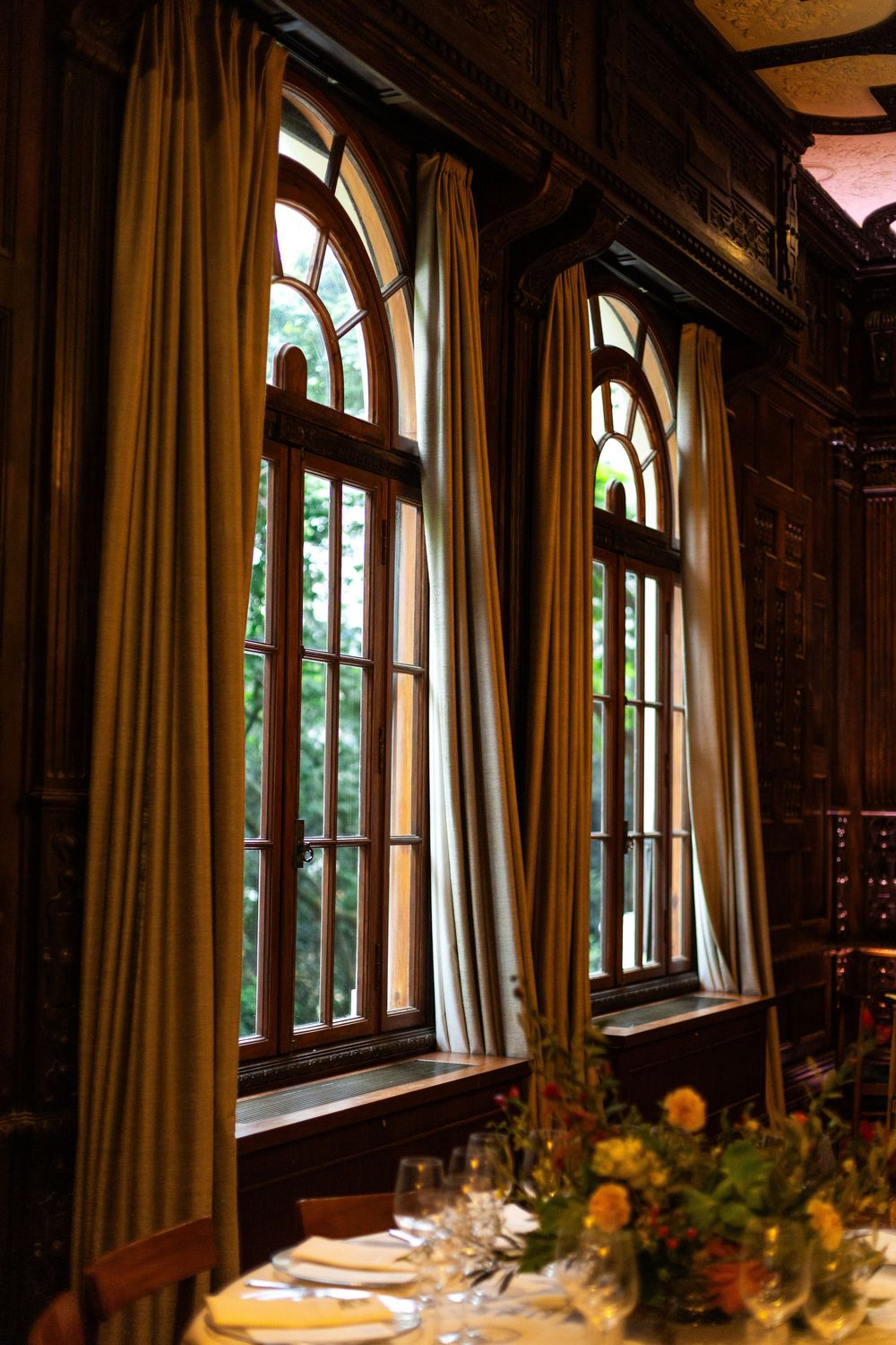 Tall arched windows with cream curtains in an ornate wood-paneled dining room.