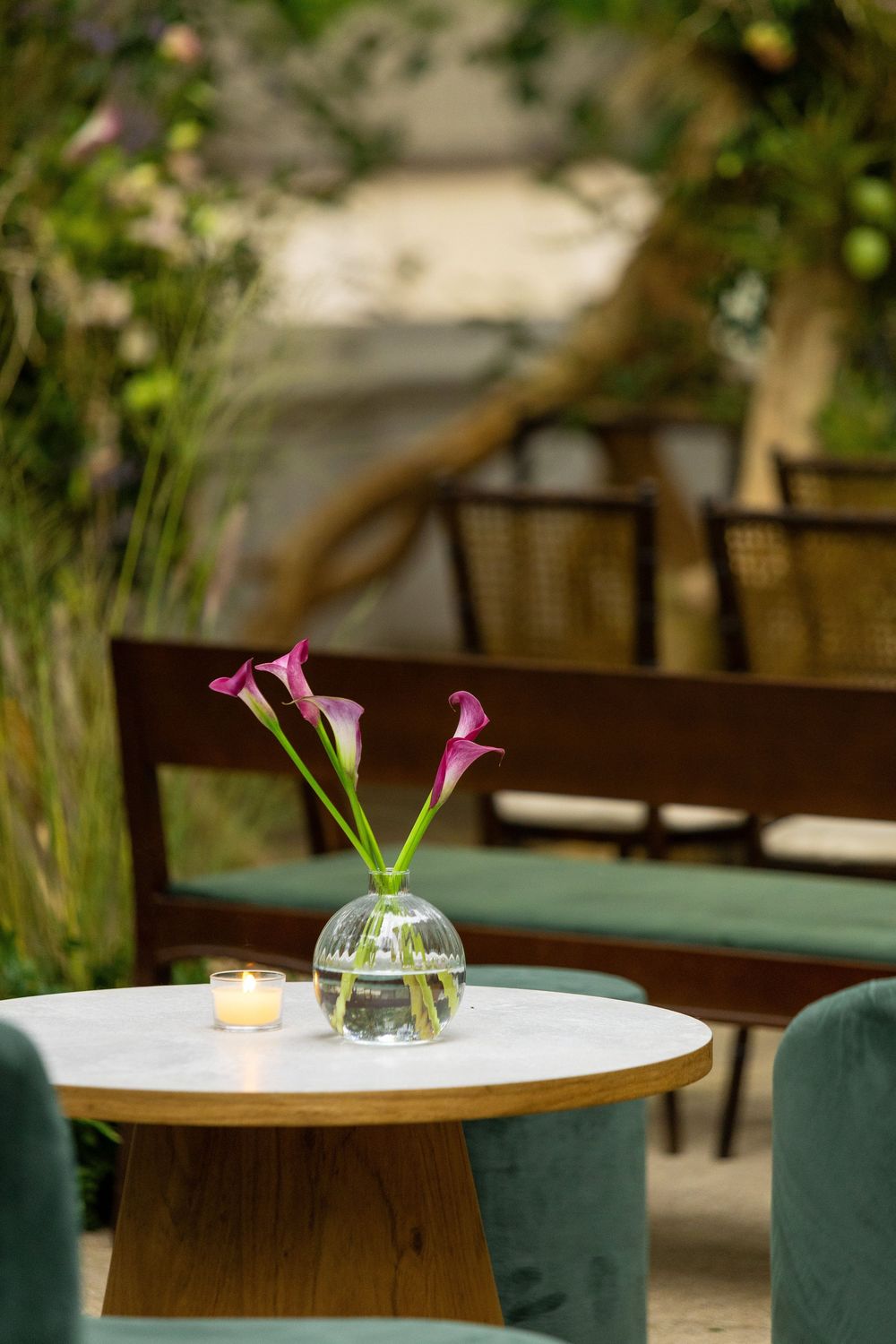 Modern restaurant tables with green velvet chairs and simple pink calla lily centerpieces in glass vases.