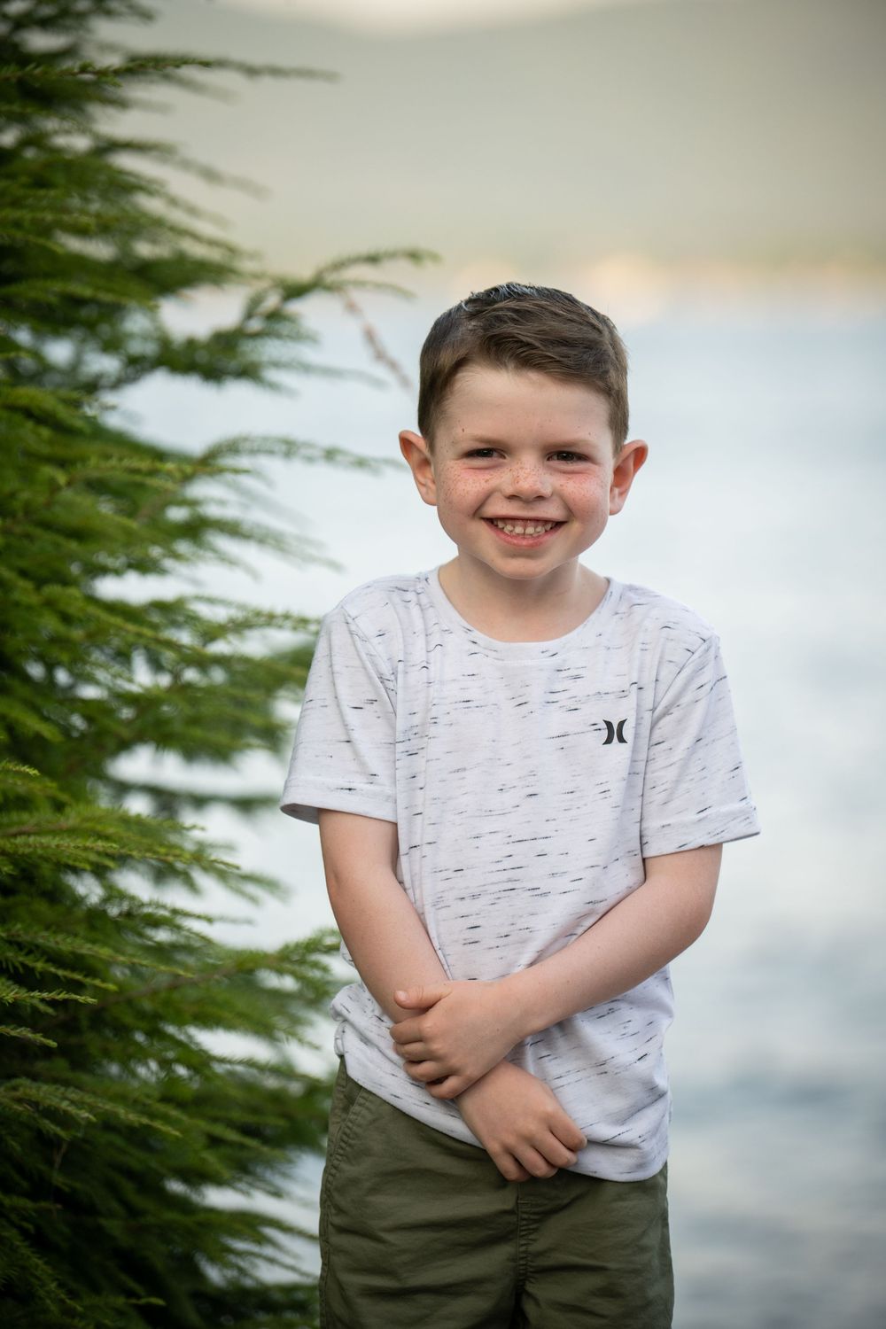 A child in a white t-shirt and olive pants poses against a blurred natural background.