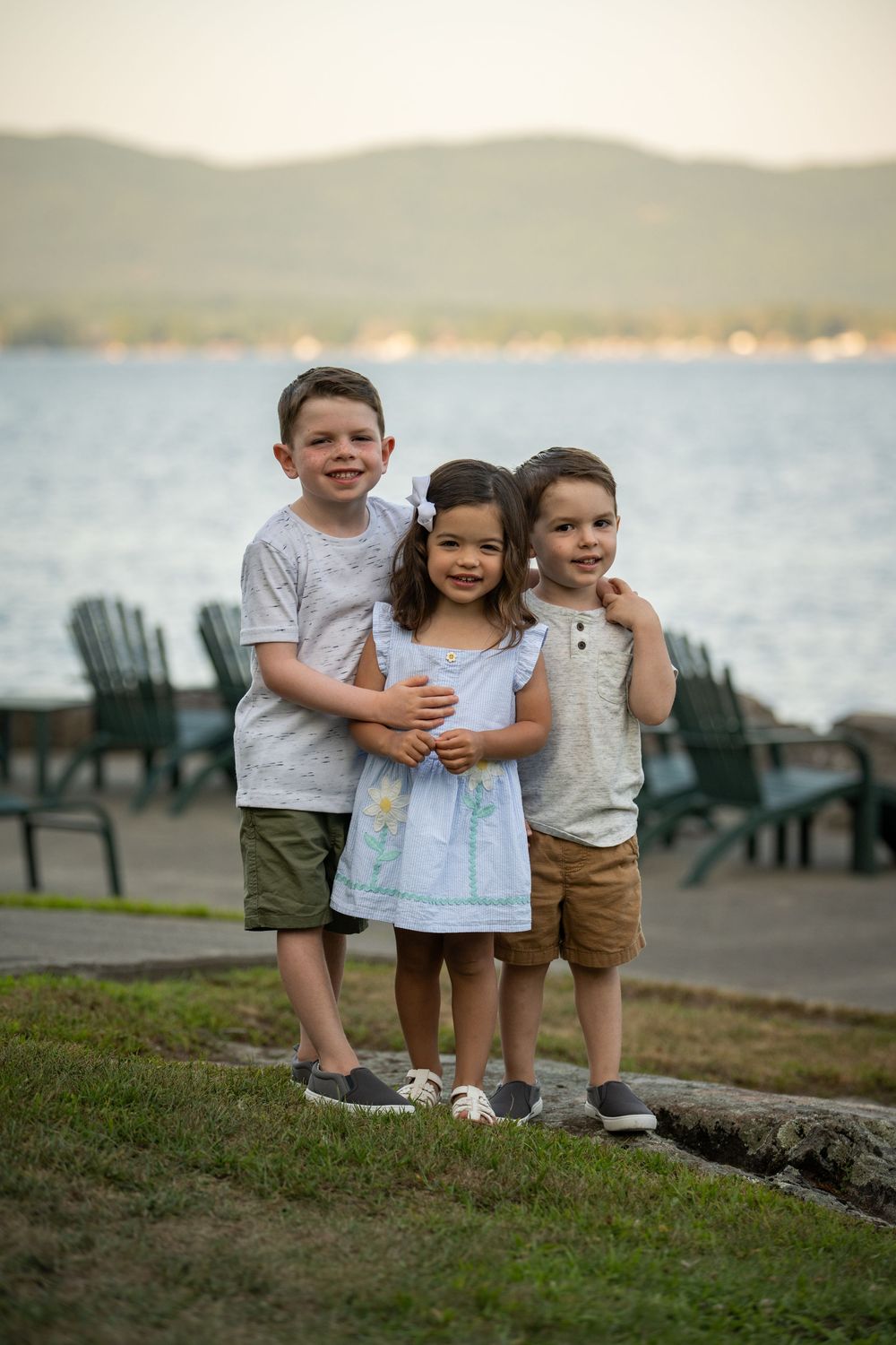 Three small children stand together by the water on a grassy hill with mountains in the background.