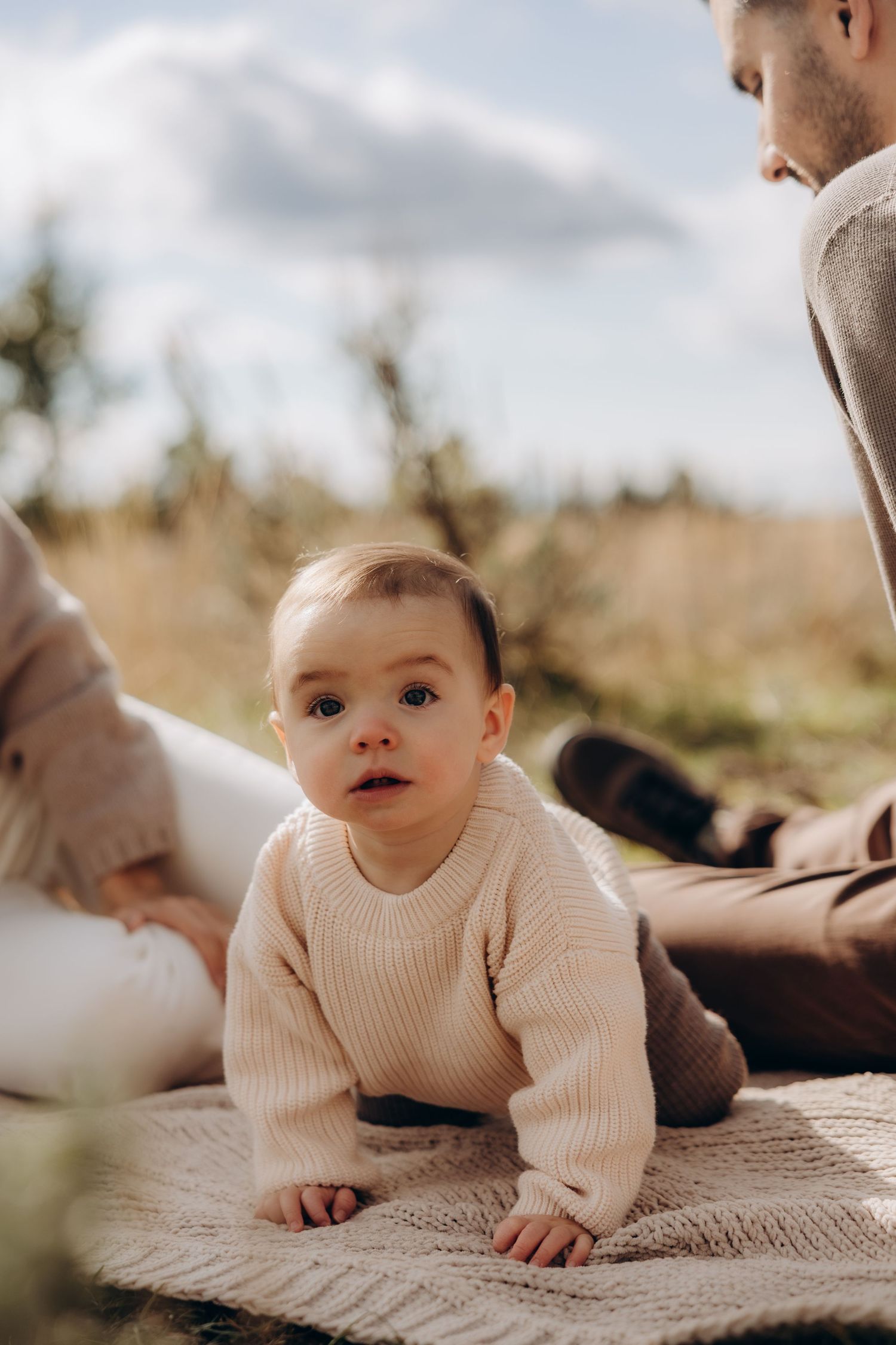 Baby wearing cream knit sweater lying on textured blanket in outdoor setting with soft natural lighting.