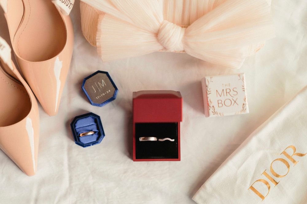 Engagement ring in red box surrounded by pink rose petals and scattered blue decor on white surface.
