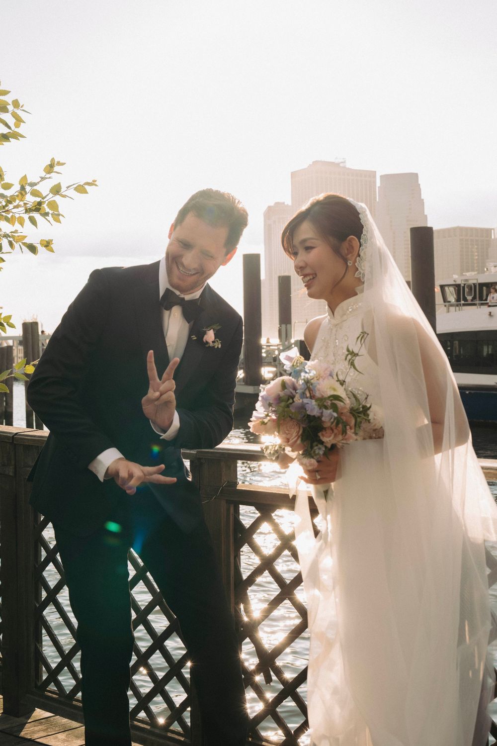 A couple in wedding attire share a playful moment on a balcony with a city skyline in the background.