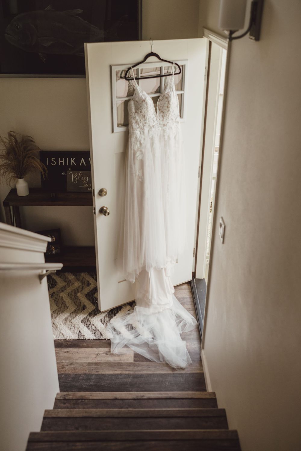 A white wedding dress hangs on a wooden hanger against a dark wall in a rustic stairwell setting.