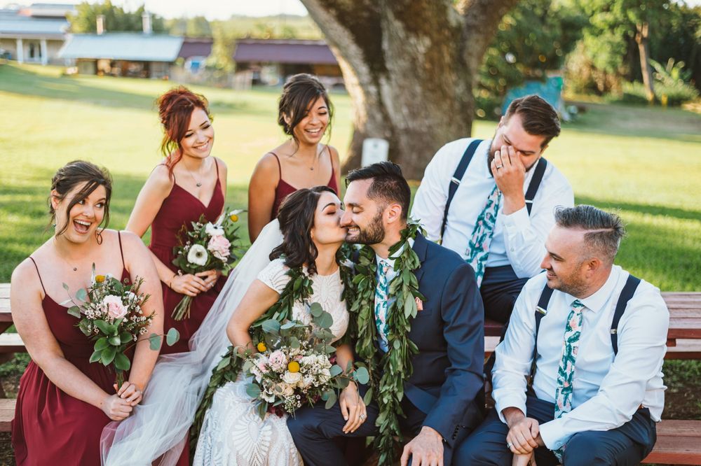 Wedding party laughs together while sitting outdoors on a sunny day in a garden setting.