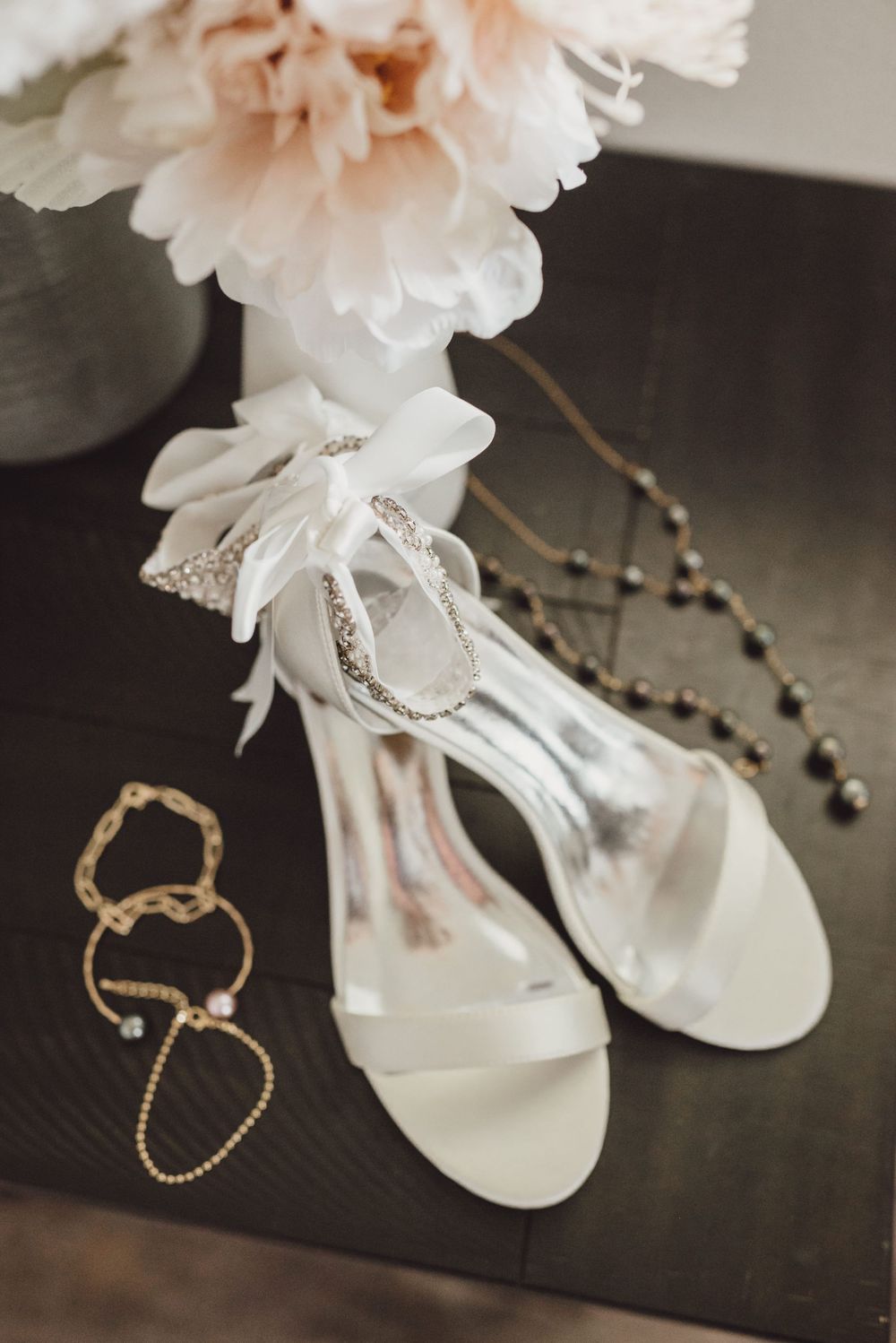Elegant bridal accessories including white sandals, delicate jewelry, and soft pink flowers arranged on dark surface.