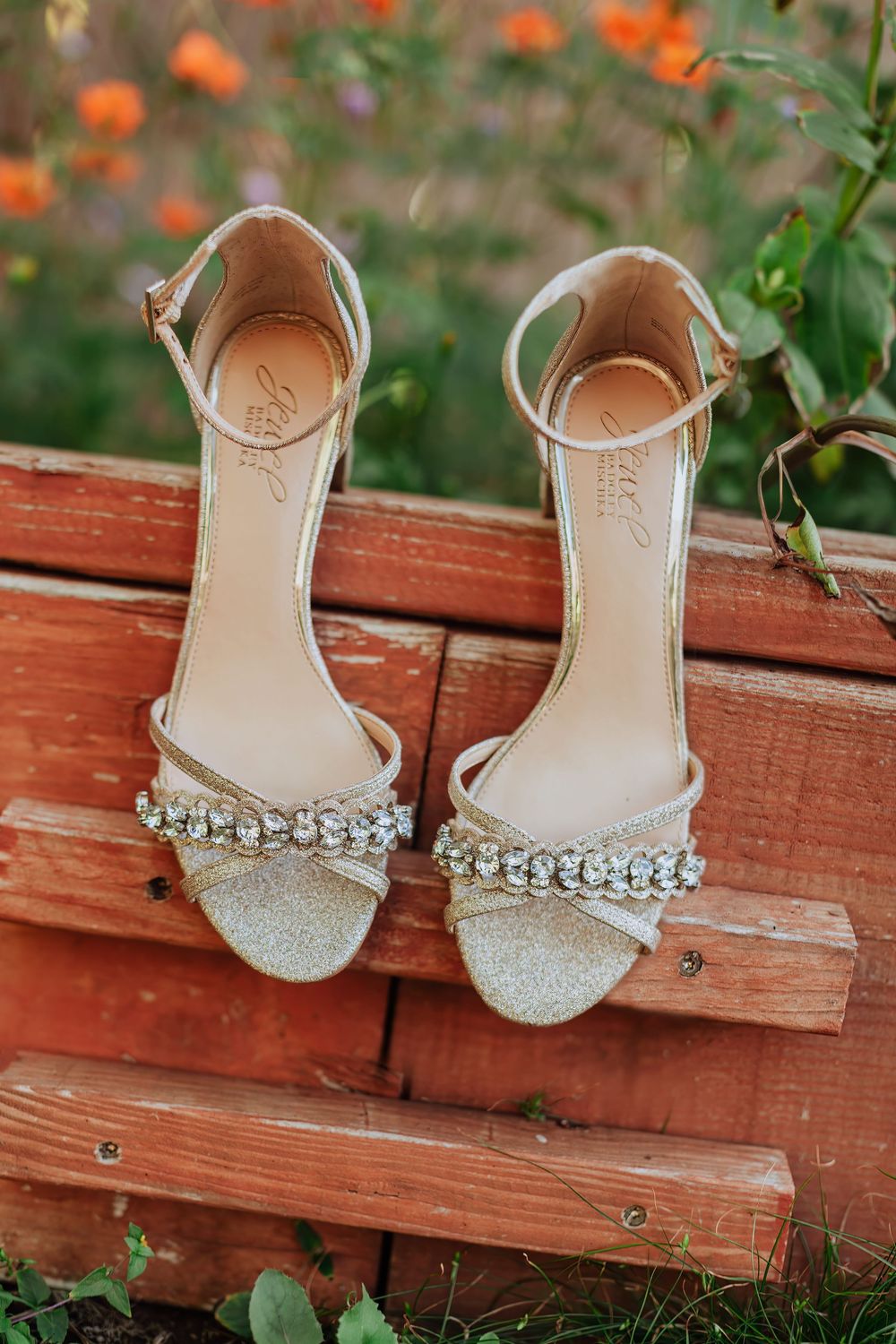 Elegant silver bridal flats with rhinestone embellishments displayed on rustic wooden deck with orange flowers in background.