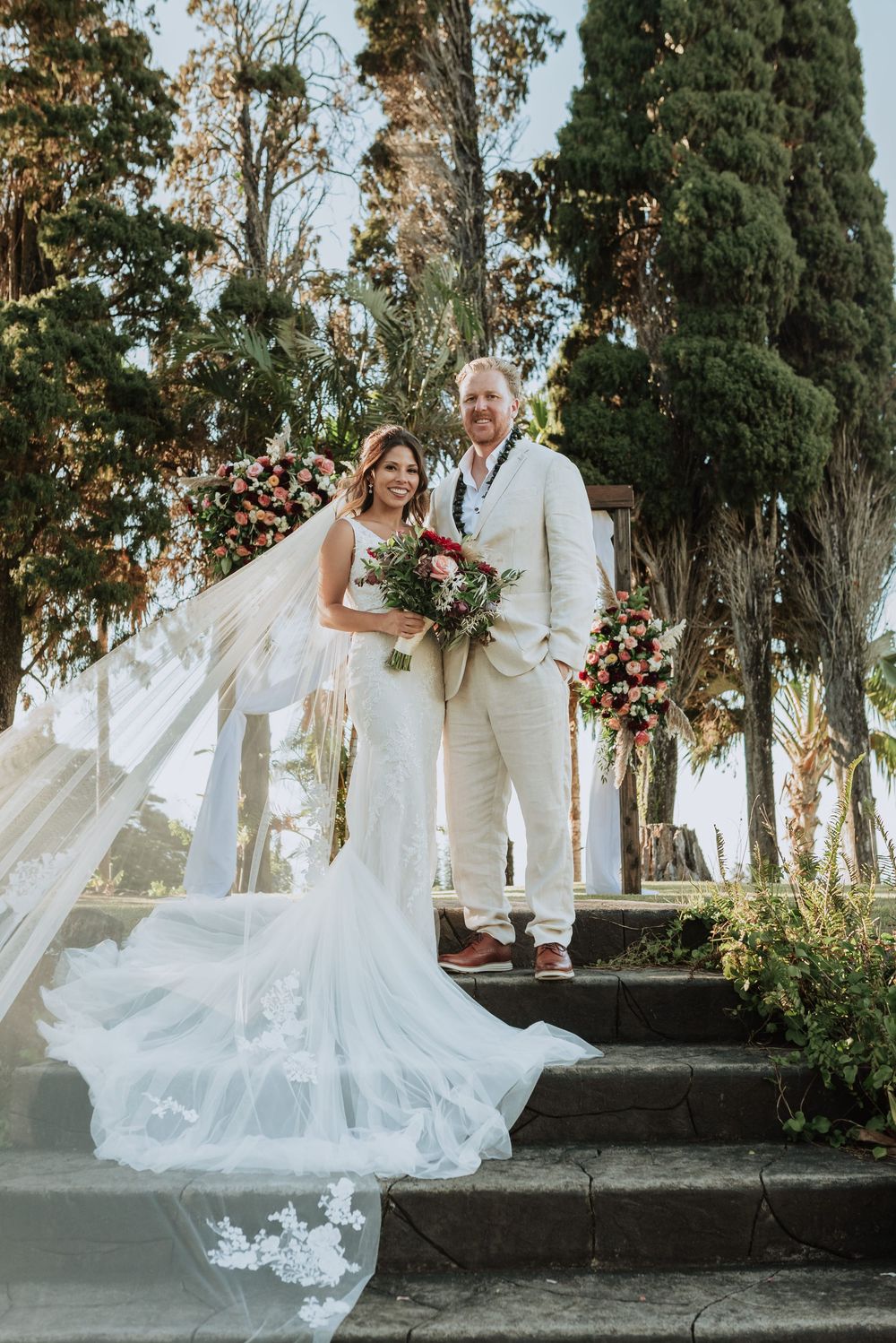 A romantic wedding couple poses on stone steps surrounded by towering cypress trees in a Mediterranean setting.