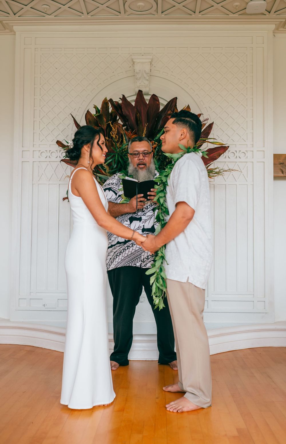 Wedding couple shares intimate first dance moment in elegant white-walled ballroom with hardwood floors and greenery decor.