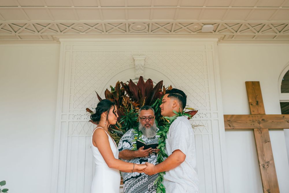 Traditional Hawaiian wedding ceremony with couple wearing green leaf leis embracing in front of white church wall.