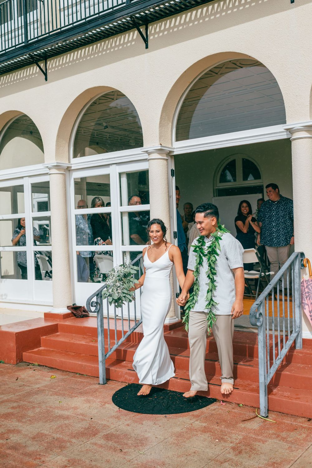 Wedding party members in white attire and green leis enter through an arched walkway at a tropical venue.