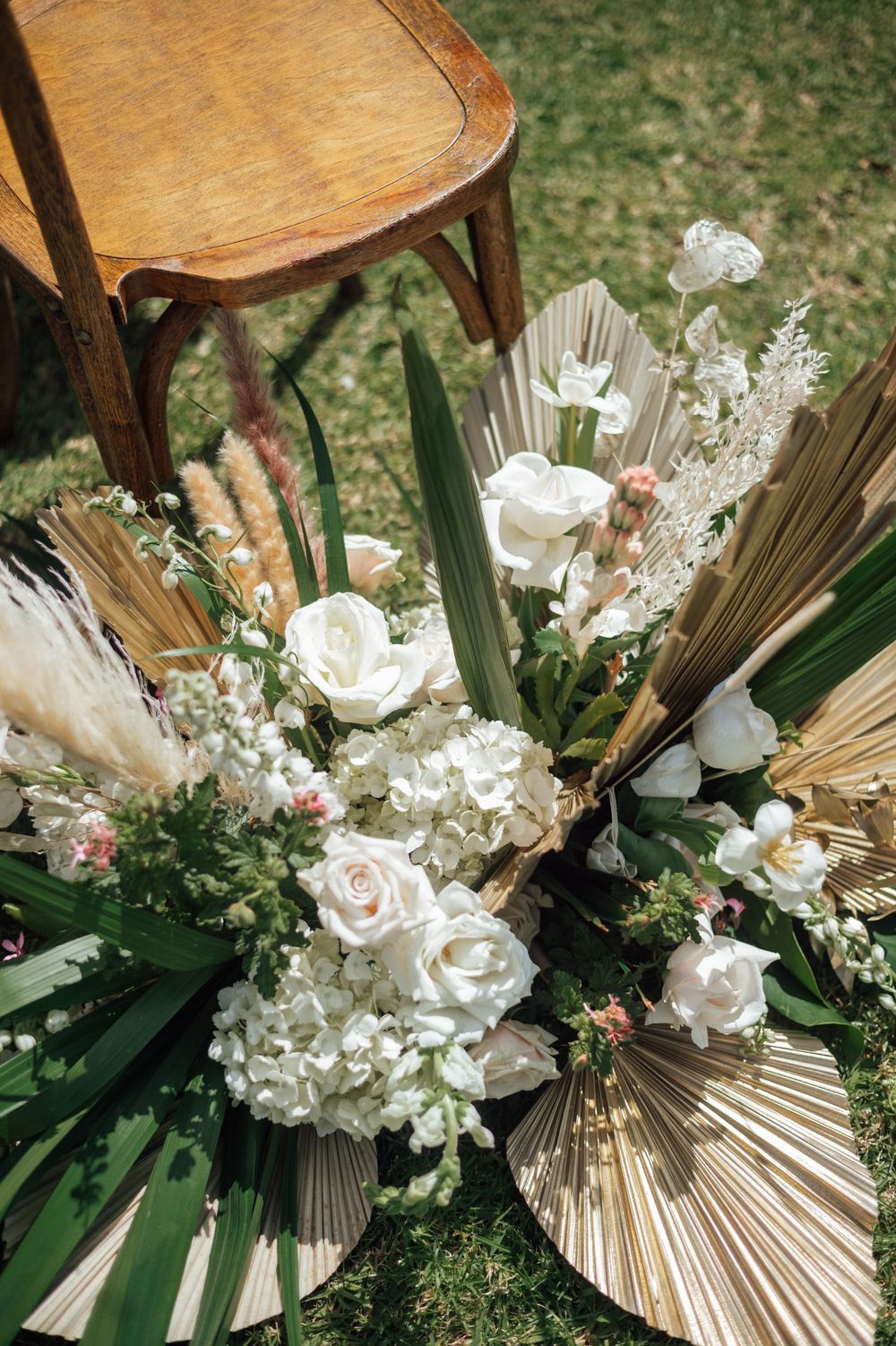 Elegant wedding floral arrangements with white roses and dried palm fronds arranged on wooden chairs outdoors.