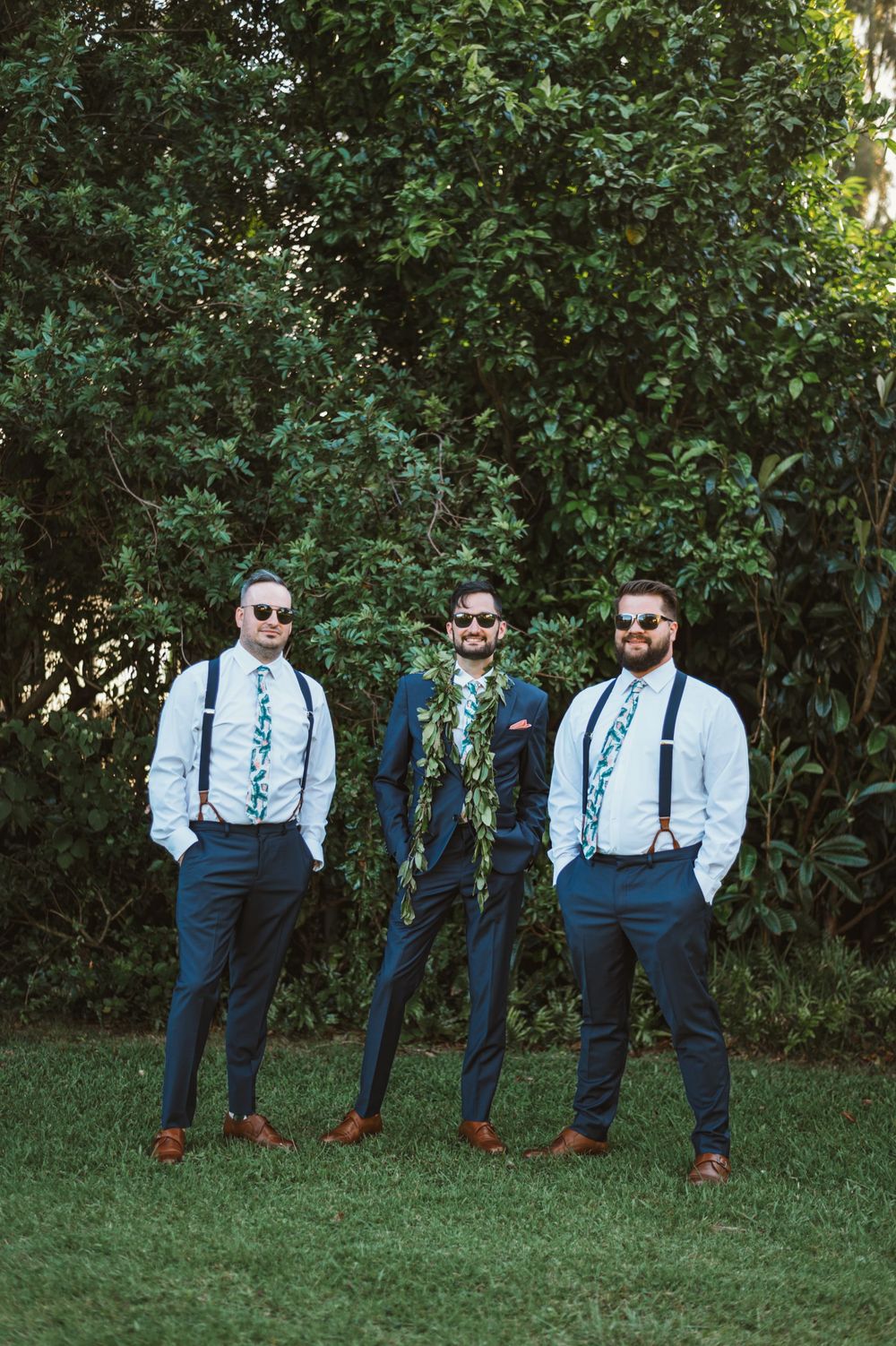 Three groomsmen in navy suits and suspenders pose casually on grass with sunglasses.