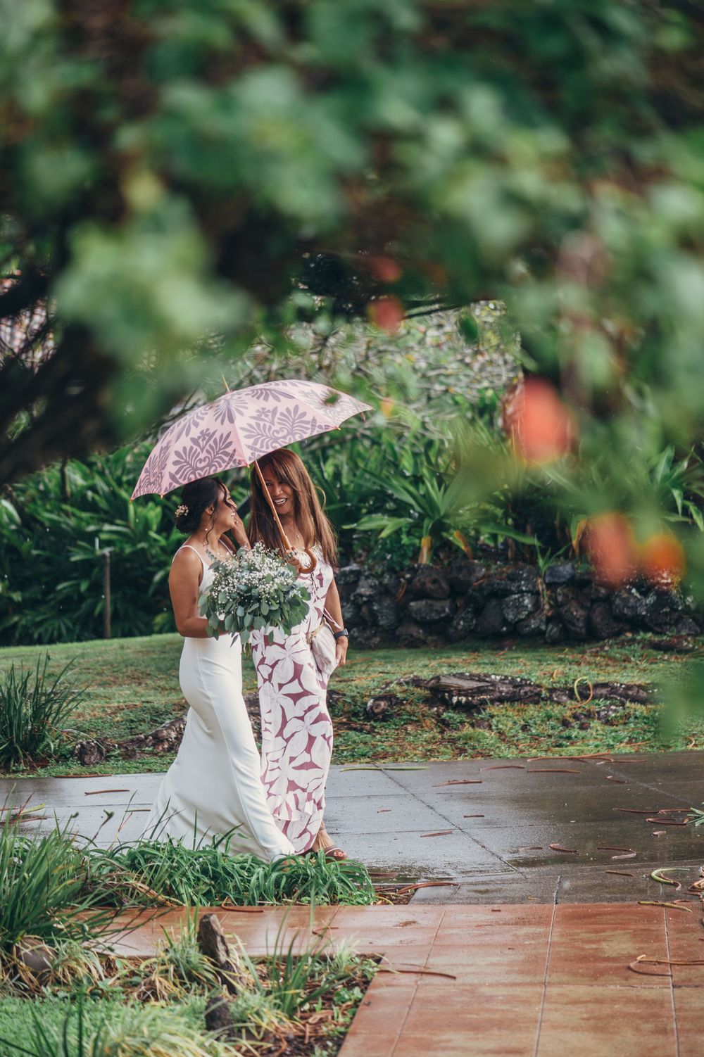 A couple shares a romantic moment under a pink umbrella on a rainy day in a garden setting.