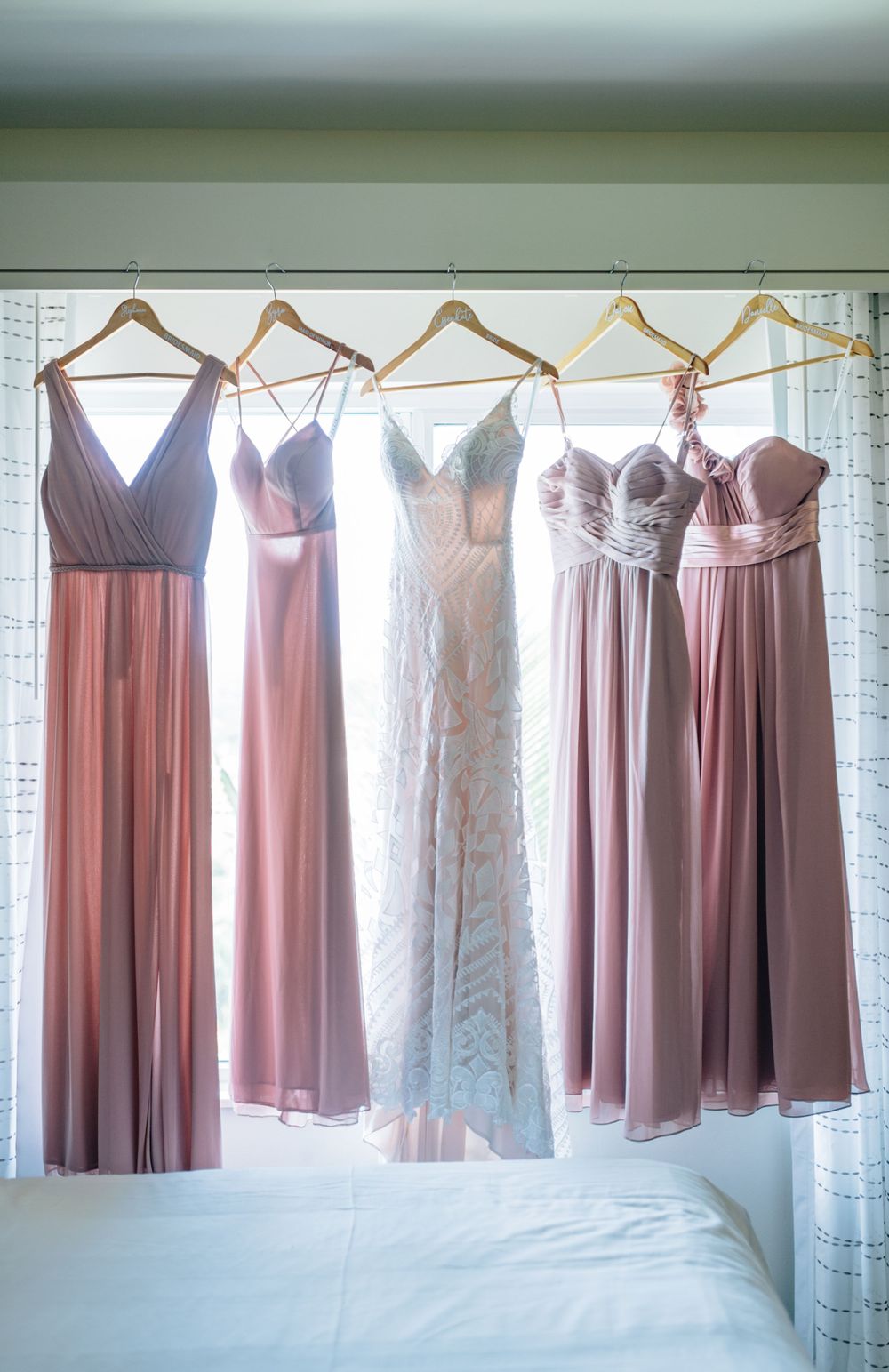 Bridesmaid dresses in various shades of pink and mauve hanging on wooden hangers against a white wall.