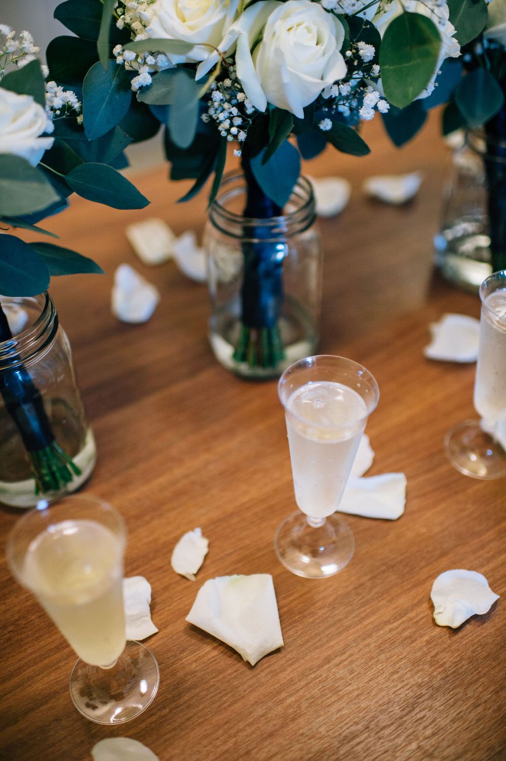 White roses and eucalyptus in mason jars with champagne glasses on wooden table.