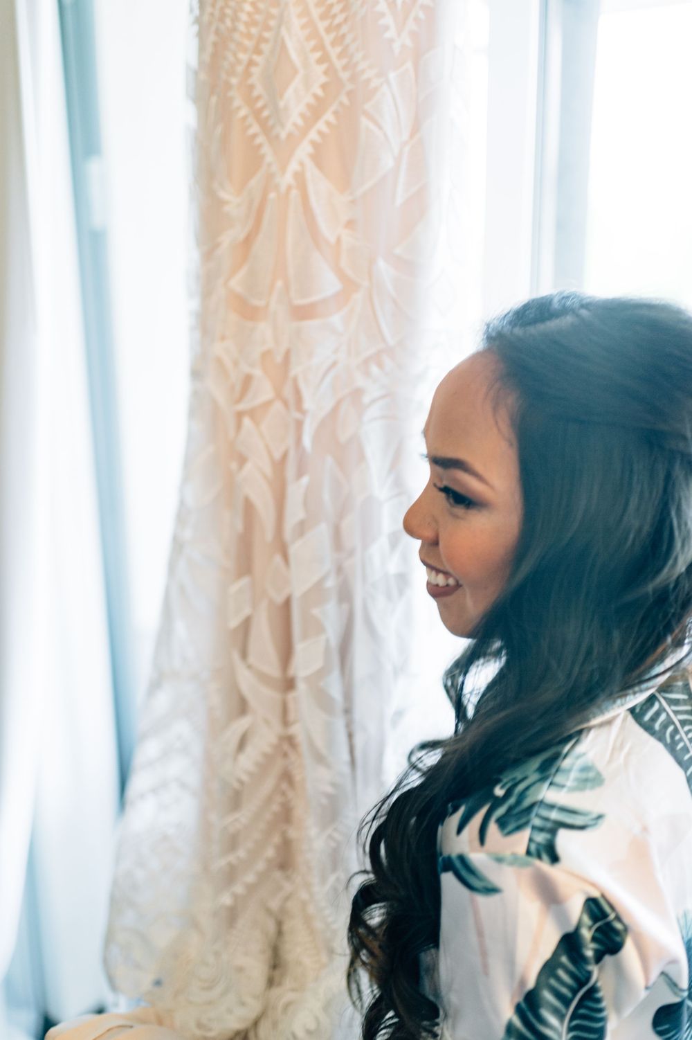 Bride in robe admiring lace wedding dress hanging by window.