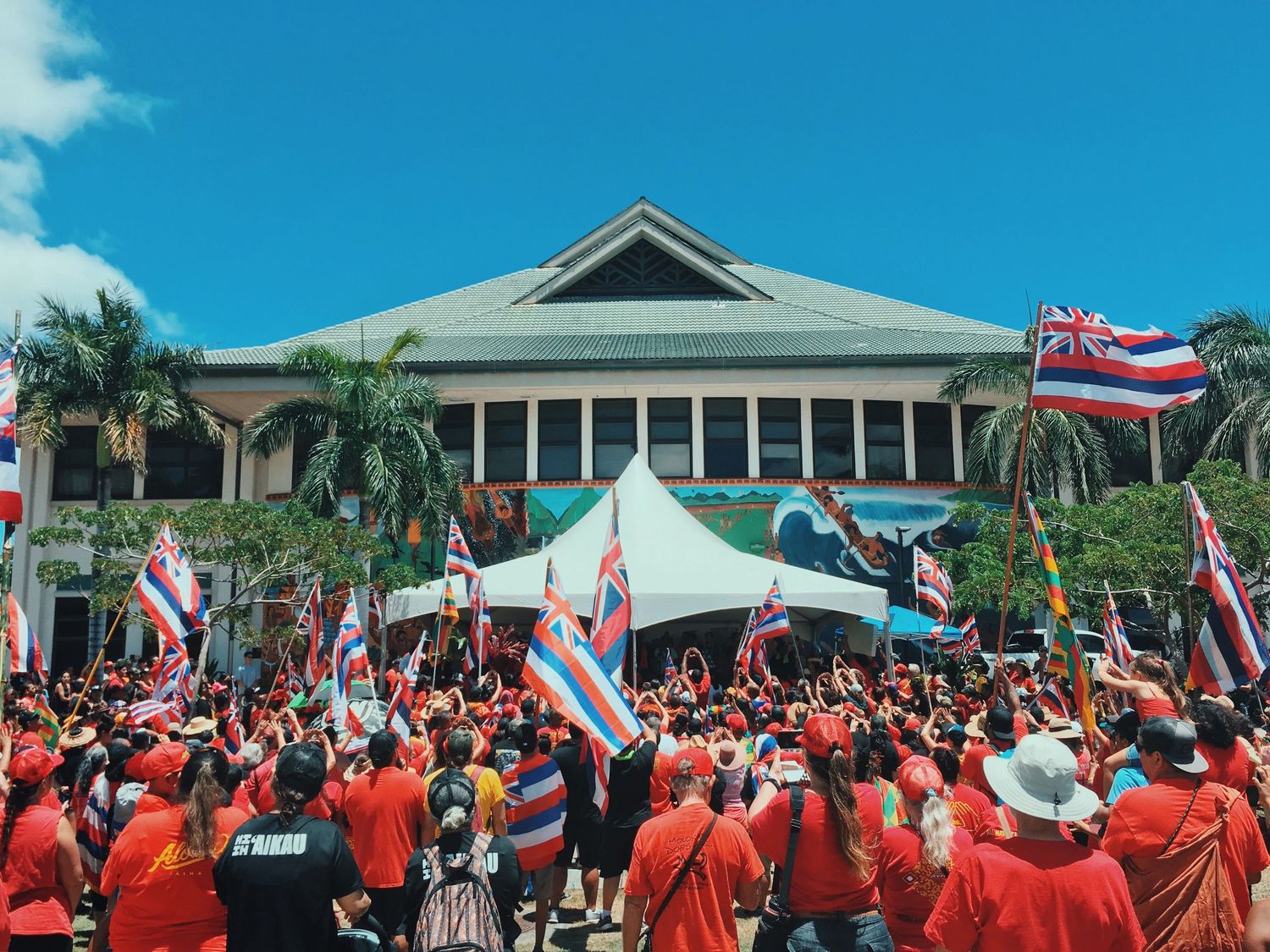 Large crowd with Hawaiian flags gathers outside buildings during daytime protest rally.
