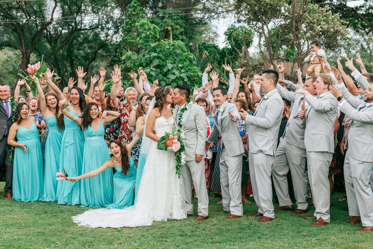 Wedding party in turquoise dresses and gray suits celebrates outdoors under tropical foliage.