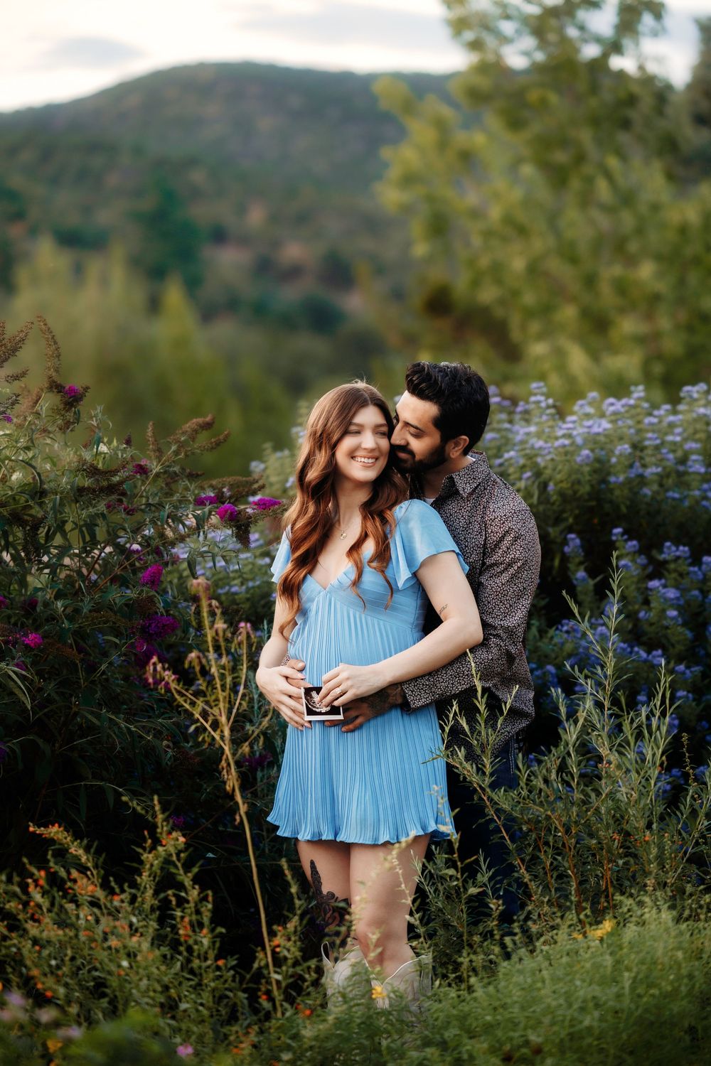 Couple sharing tender moment in flower garden, man kisses pregnant woman in blue dress holding ultrasound.