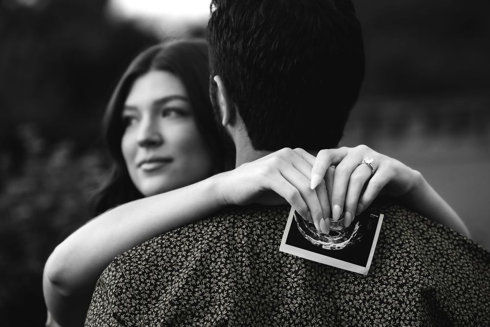 Black and white photo of woman hugging man from behind holding ultrasound photo.