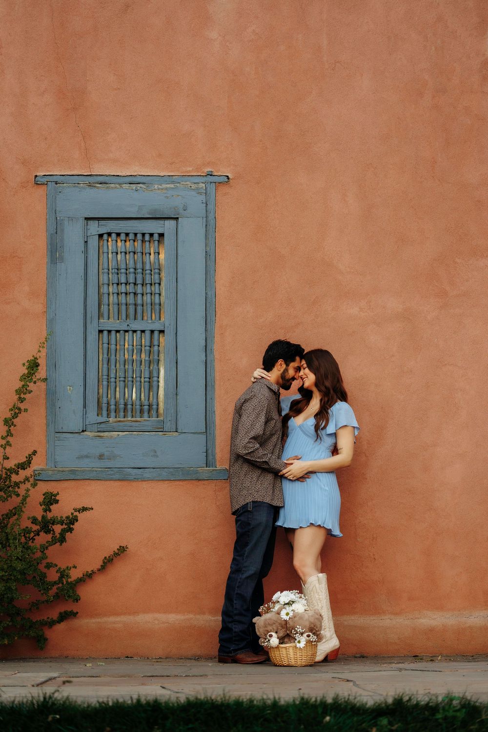 Couple kissing against terracotta adobe wall with blue window and flower basket below.