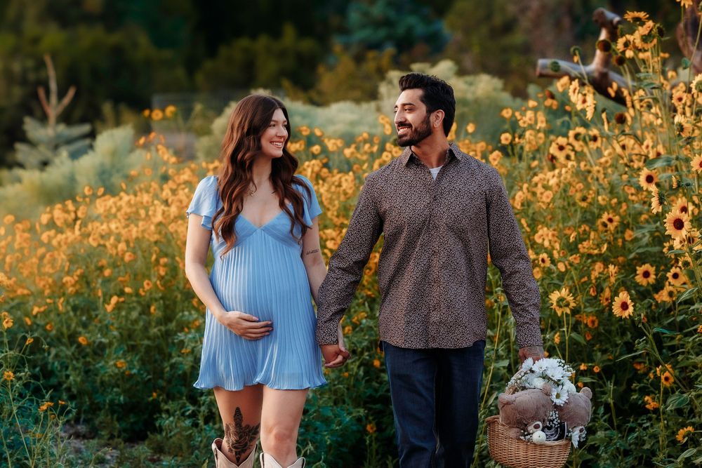 Expecting couple walks hand-in-hand through golden sunflower field, woman in blue dress and cowboy boots.
