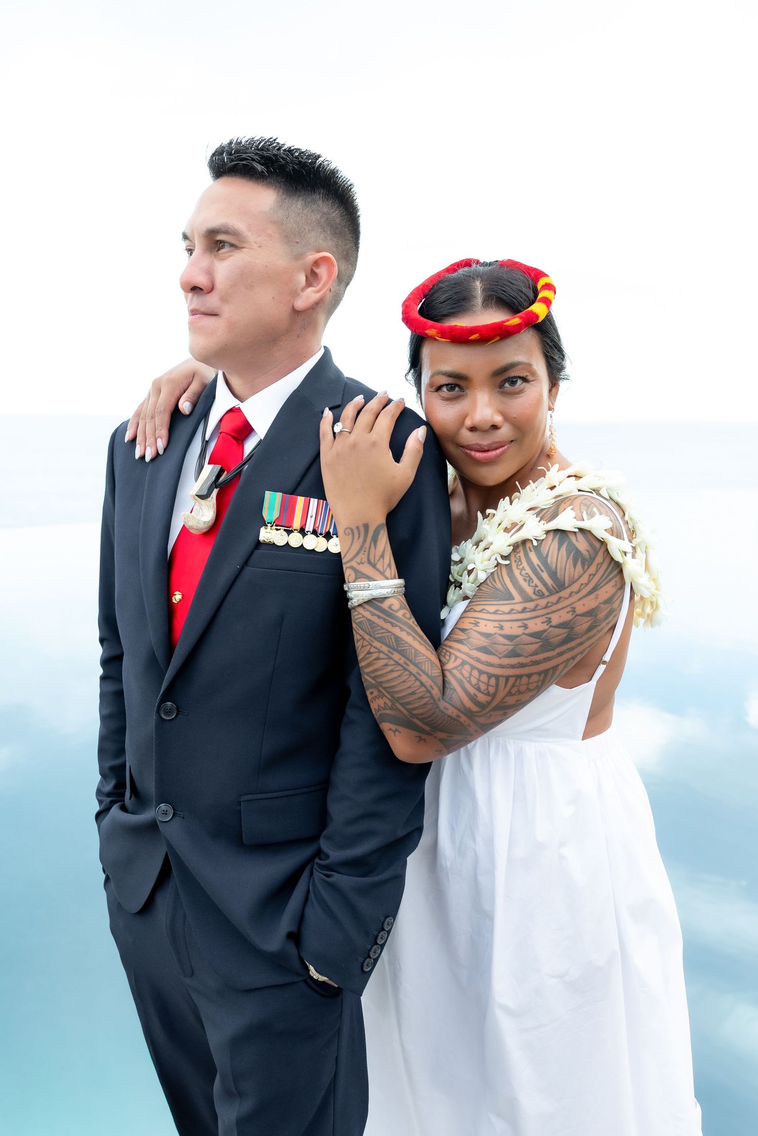 Military wedding couple embraces in formal attire against blue sky background.