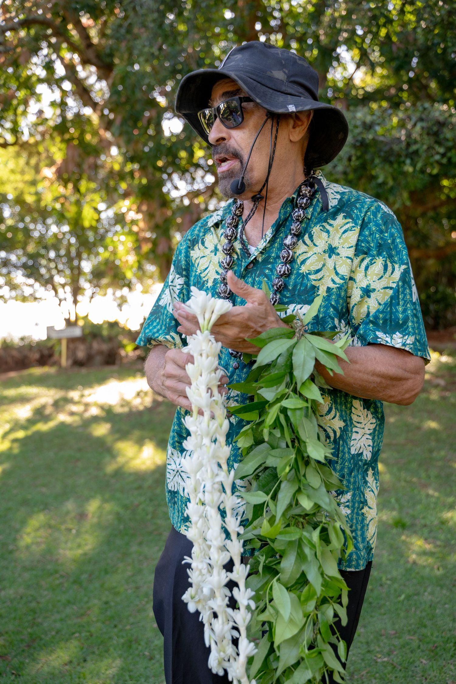 Person wearing Hawaiian shirt and hat holds white orchid lei and green foliage in outdoor garden setting.