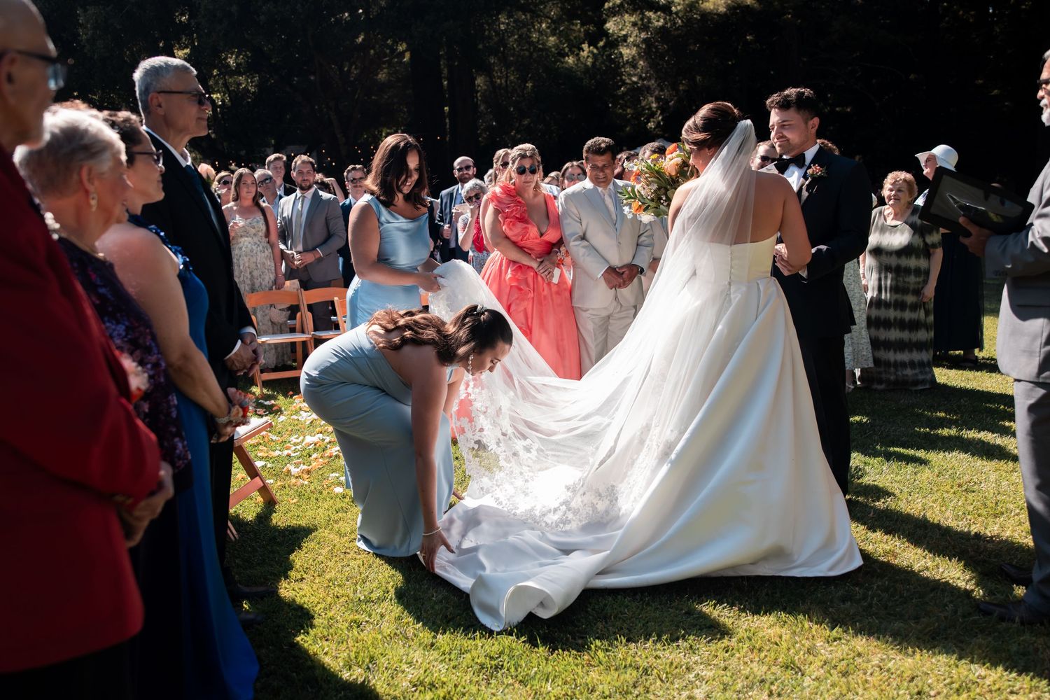 Wedding party gathers outdoors with mountains in background as bride's white dress flows elegantly in breeze.
