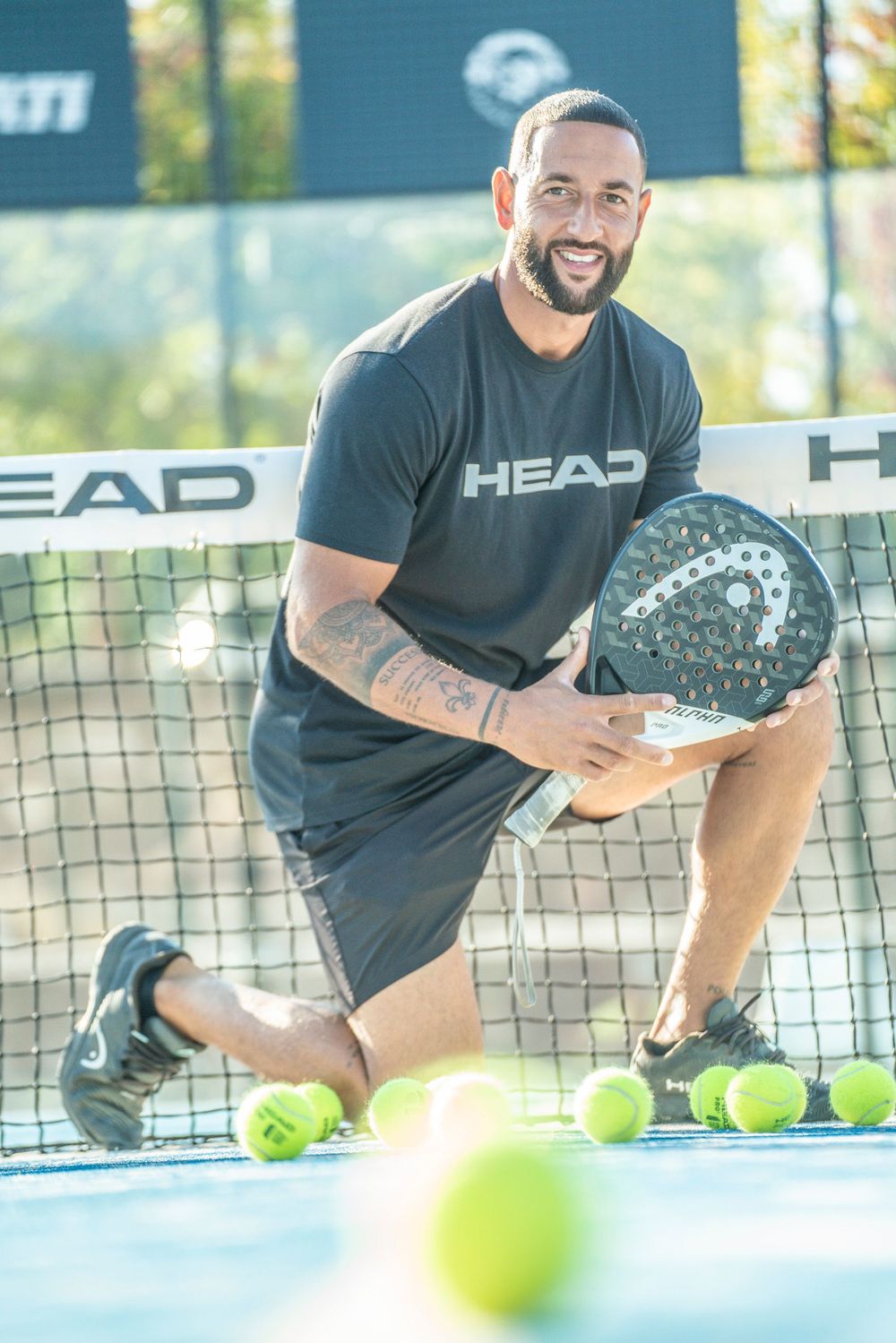 Tennis player in HEAD branded apparel kneels on court with paddle and tennis balls during outdoor training session.