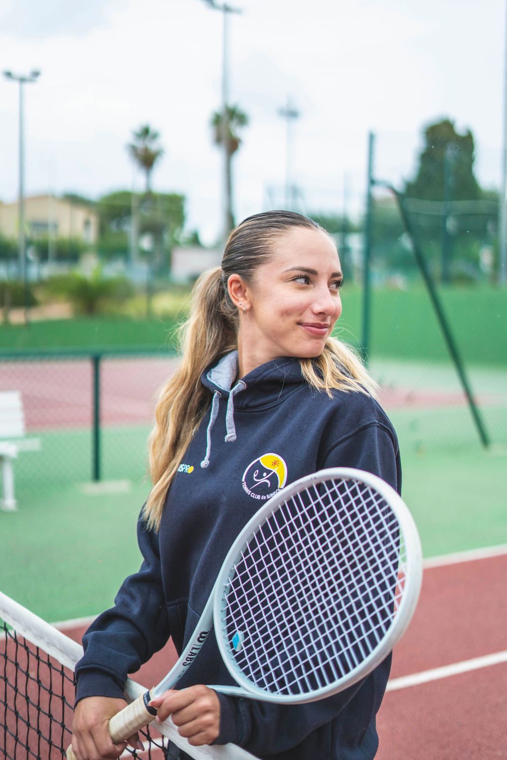 Tennis player in navy jacket holds racquet while standing on outdoor court with palm trees in background.