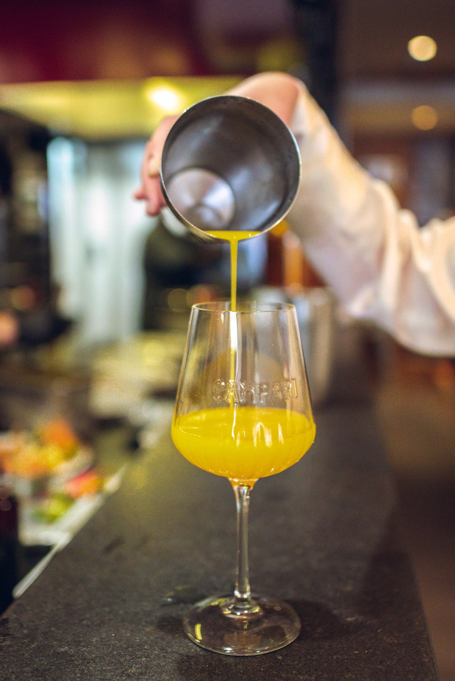 A golden-colored cocktail being poured from a mixing glass into an elegant stemmed glass at a dimly lit bar.