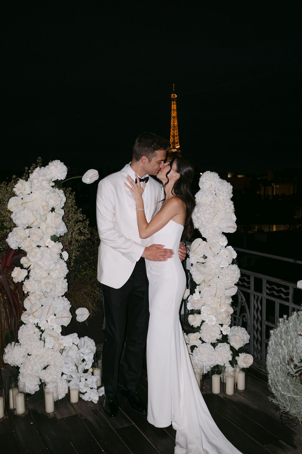 Romantic wedding couple shares a kiss under white floral arch with illuminated Eiffel Tower in background at night.