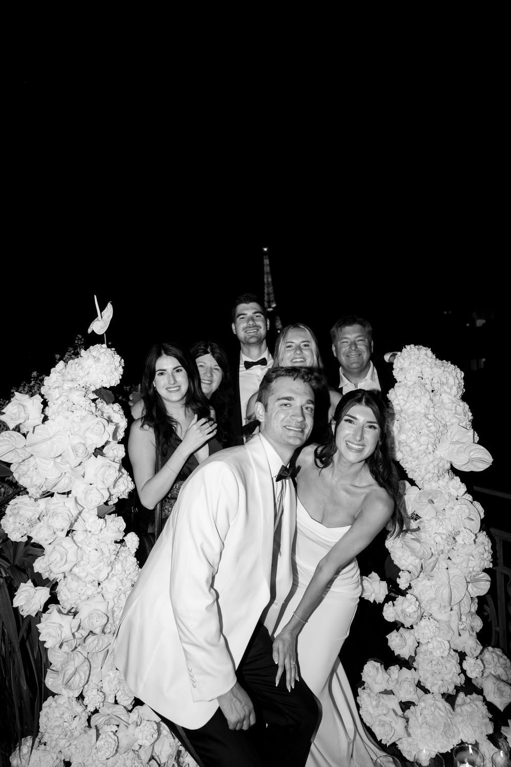 A group poses together in a black and white photo with decorative white floral arrangements.