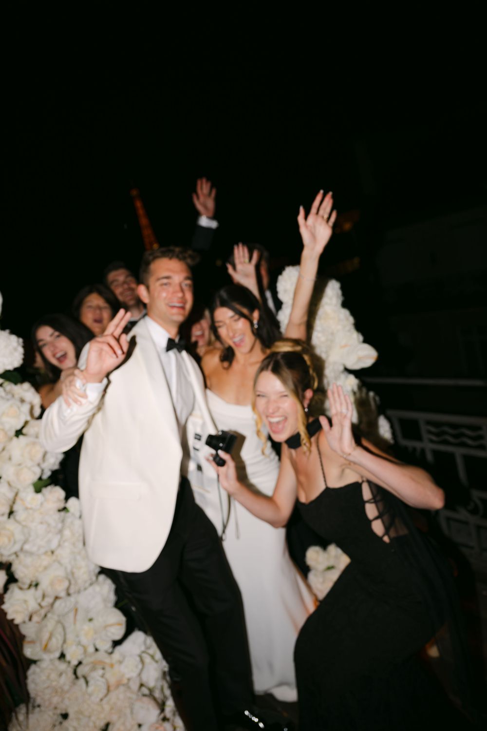 Wedding celebration with guests dancing and cheering amid white floral decorations at a nighttime reception.
