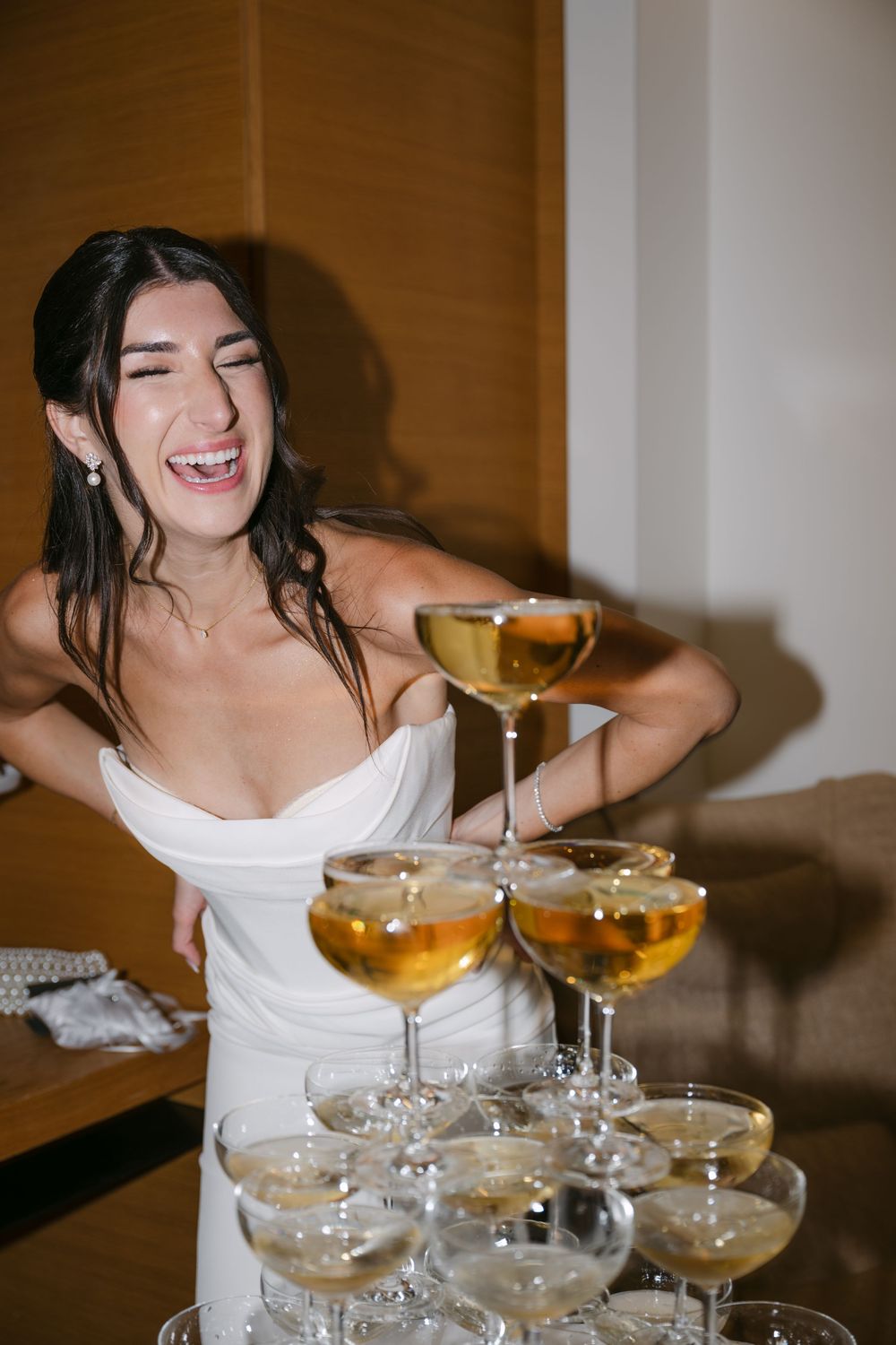 Bride in white dress laughs while posing with champagne tower at elegant wedding reception.