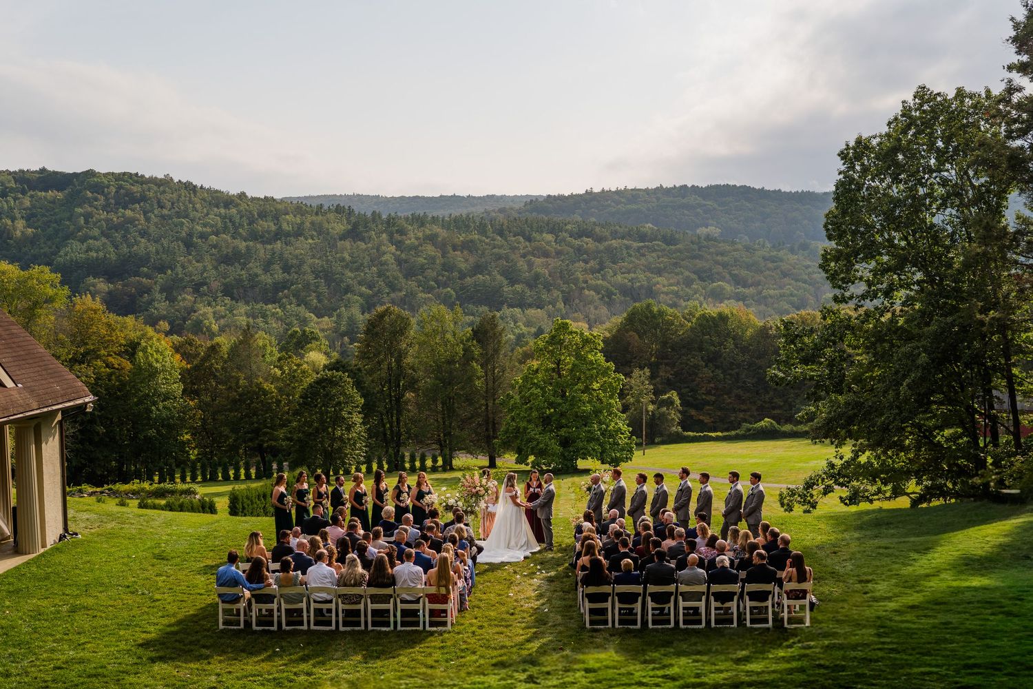 A panoramic view of an elegant outdoor wedding ceremony setup with rows of chairs on a lush green lawn surrounded by mountains.