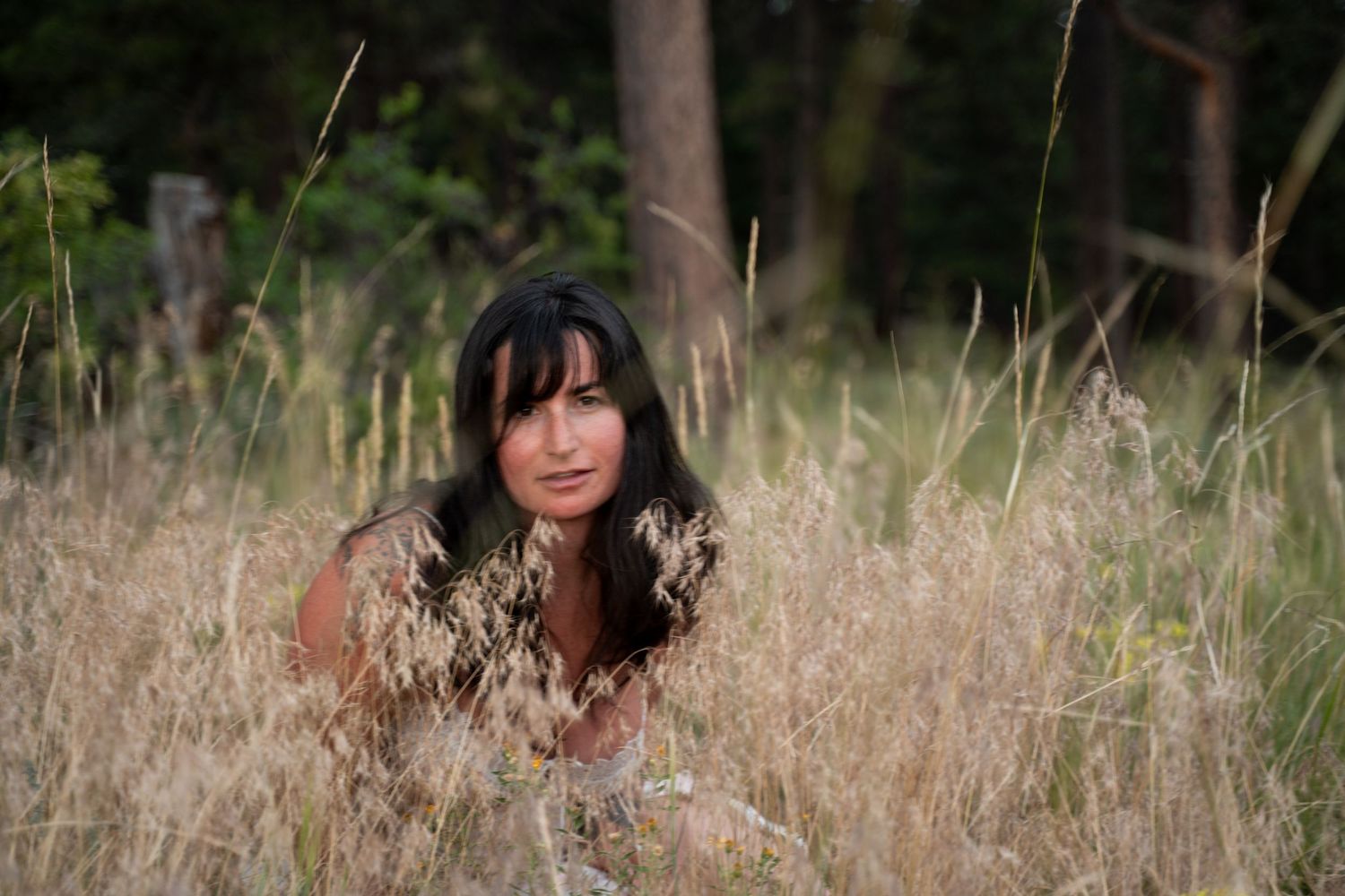 A person with dark hair poses thoughtfully in a field of tall dried grass and autumn foliage.