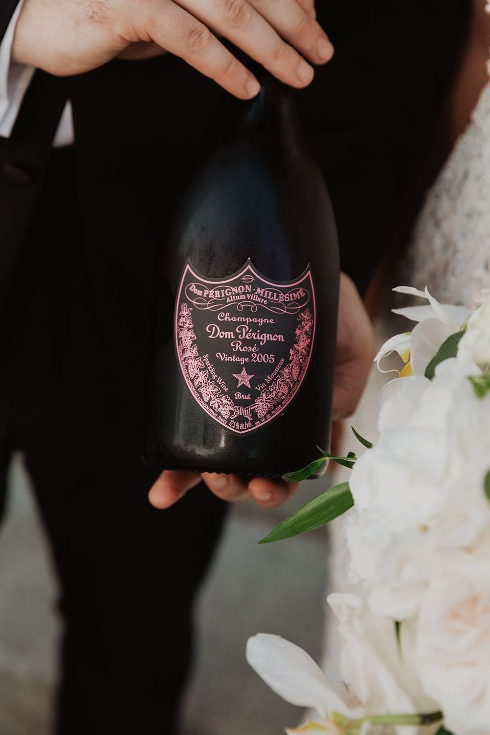 Couple holds Dom Perignon Rose Vintage 2005 champagne bottle beside white orchids at wedding.