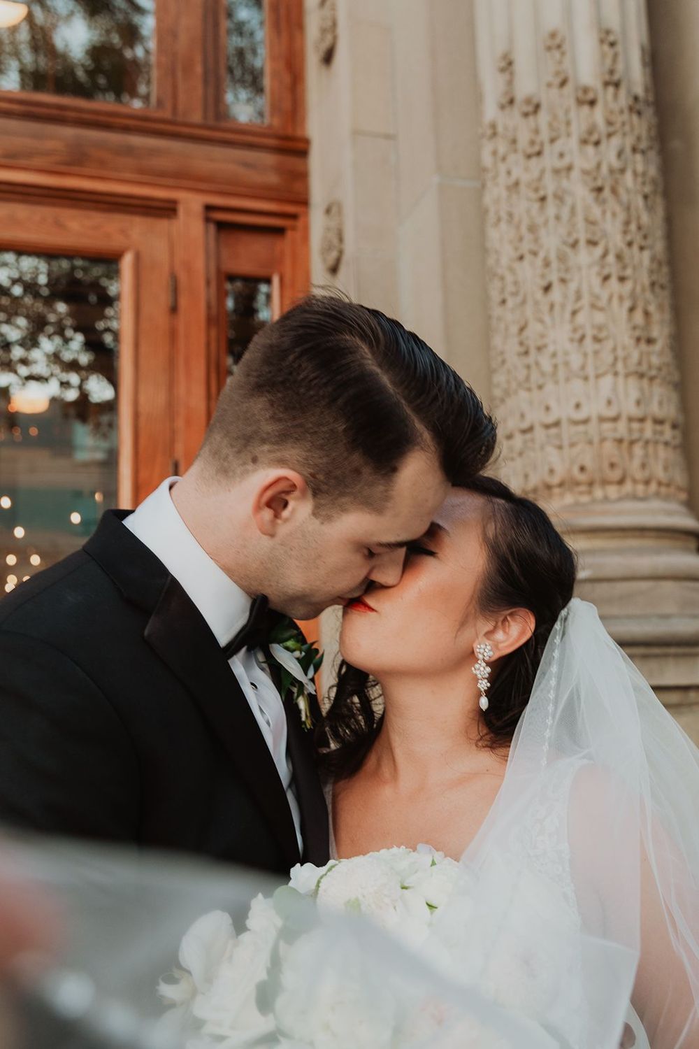 Bride and groom share a romantic kiss outside ornate stone building with wooden doors at sunset.