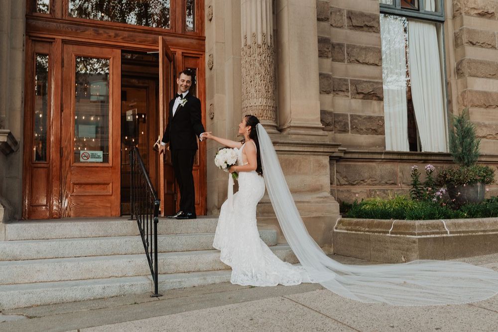 Bride in mermaid gown holds grooms hand on steps of elegant stone building with wooden doors.