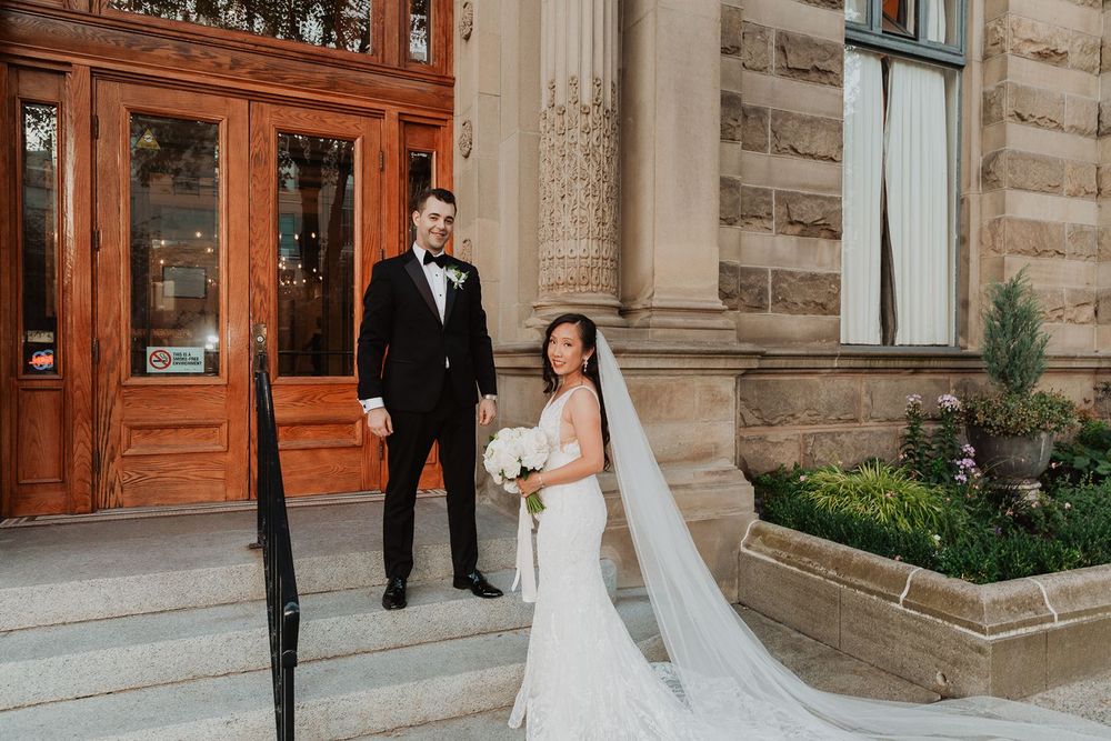 Bride in fitted gown with cathedral veil and groom in tuxedo pose outside historic stone venue.