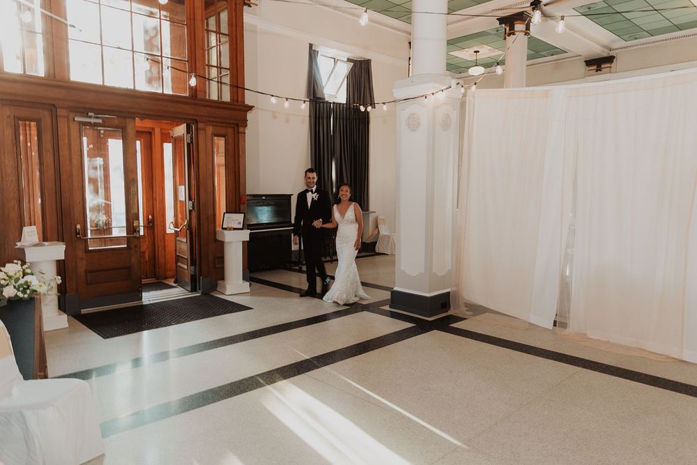 Bride and groom stand in grand marble foyer of elegant wedding venue with string lights above.