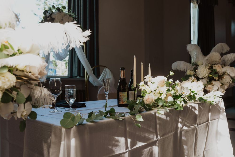 Elegant wedding sweetheart table with white floral arrangements, feathers, candles, and champagne bottle.