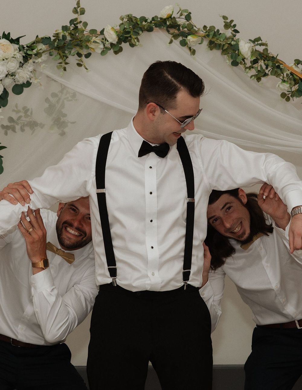 Groom in bow tie and suspenders posing playfully with two groomsmen at wedding arch.