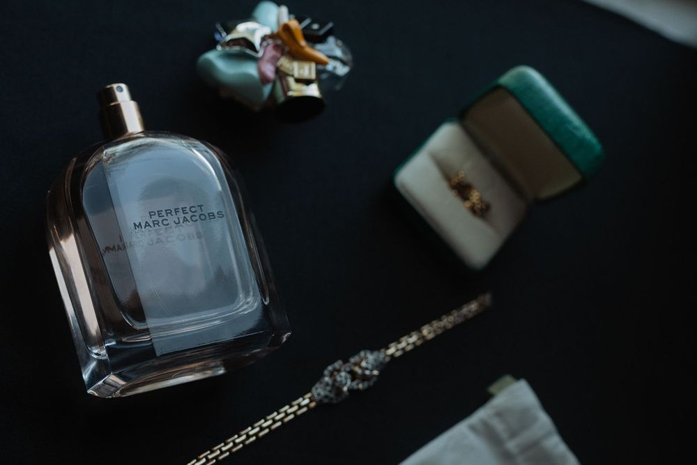 Marc Jacobs Perfect perfume bottle displayed with wedding rings and jewelry on dark surface.